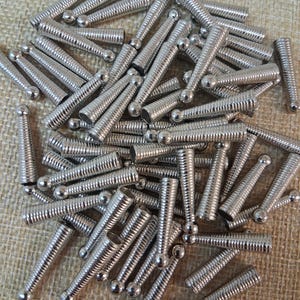 Op de afbeelding: Een stapel kleine, zilverkleurige metalen schroefbevestigingen. Elke bevestiger heeft een cilindrisch lichaam met een spiraalvormige schroefdraad en een afgeronde kop. De bevestigingen zijn verspreid over een getextureerd, beige oppervlak.
