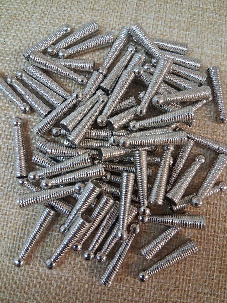 Op de afbeelding: Een stapel kleine, zilverkleurige metalen schroefbevestigingen. Elke bevestiger heeft een cilindrisch lichaam met een spiraalvormige schroefdraad en een afgeronde kop. De bevestigingen zijn verspreid over een getextureerd, beige oppervlak.