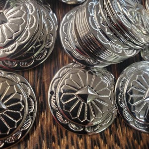 Puede incluir: Primer plano de varios conchos de metal plateado brillante. Cada concho tiene un diseño detallado con una forma de estrella central y un borde decorativo. Los conchos están apilados y dispersos, reflejando la luz.