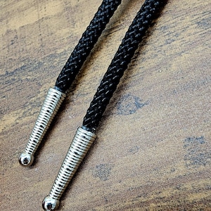 Op de afbeelding: Een zwarte bolo stropdas met een zilverkleurige cirkelvormige schuif en decoratieve uiteinden. Het koord van de stropdas is zwart en gevlochten. De uiteinden zijn zilverkleurig en hebben een geribbeld ontwerp.