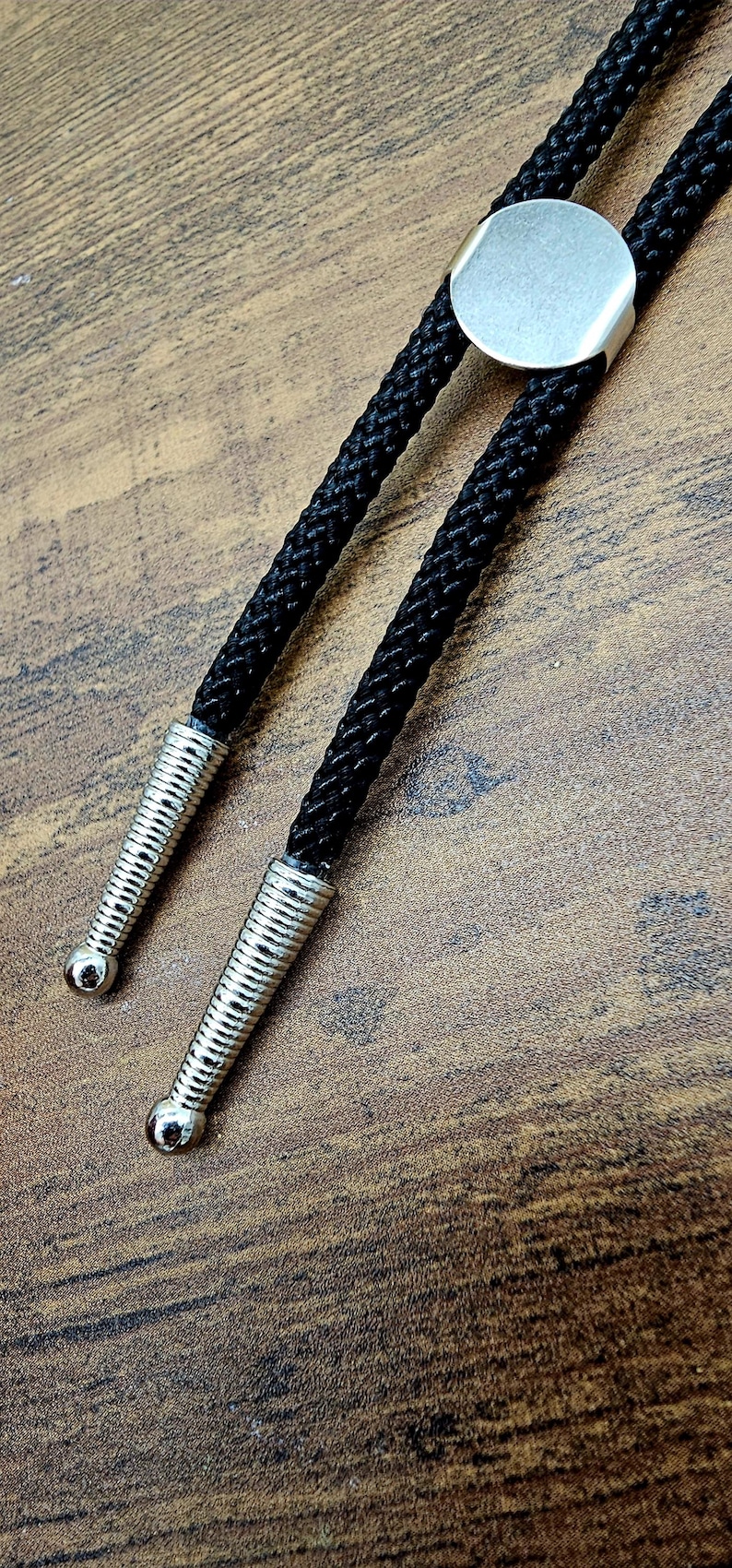 Op de afbeelding: Een zwarte bolo stropdas met een zilverkleurige cirkelvormige schuif en decoratieve uiteinden. Het koord van de stropdas is zwart en gevlochten. De uiteinden zijn zilverkleurig en hebben een geribbeld ontwerp.