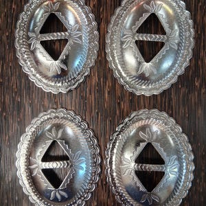 May include: Four oval-shaped silver-toned metal concho buckles featuring a scalloped edge, a central diamond-shaped cutout with a horizontal bar, and engraved leaf-like patterns surrounding the centre. Each buckle has a polished finish.