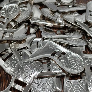 May include: A pile of shiny silver-colored metal belt buckles. The buckles have an ornate design with floral and star patterns. The buckles are in a variety of shapes and sizes, with a slot for the belt to go through.