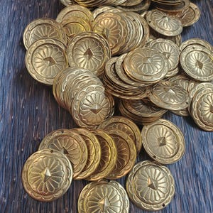 Puede incluir: Una gran pila de adornos redondos decorativos de metal dorado con un diseño geométrico en relieve. Los adornos tienen un borde festoneado y un diseño central en relieve.