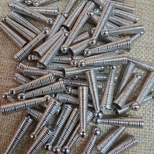 May include: A pile of silver-colored metal springs with a ball at one end. The springs have a ridged, coiled design. The objects are scattered on a textured, tan surface.