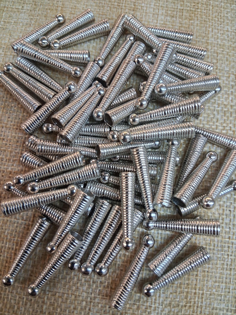 Op de afbeelding: Een stapel zilverkleurige metalen veren met een bol aan &eacute;&eacute;n uiteinde. De veren hebben een geribbeld, opgerold ontwerp. De objecten liggen verspreid op een getextureerd, bruinachtig oppervlak.
