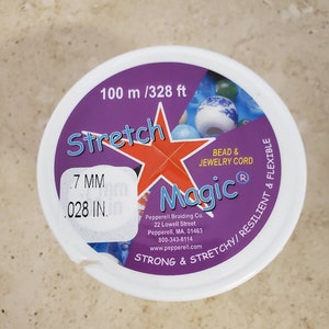 May include: A purple and white container of Stretch Magic bead and jewellery cord. The container is labelled with the text "Stretch Magic", "Bead & Jewellery Cord", "Strong & Stretchy/Resilient & Flexible", "100 m / 328 ft", and "7 MM .028 IN."