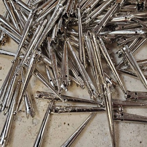 May include: A pile of silver-toned metal tips with a decorative design. The tips are pointed and have a small ball at the end.