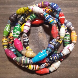 Op de afbeelding: Een kleurrijke ketting gemaakt van gerolde papier kralen. De kralen zijn in verschillende kleuren, waaronder rood, blauw, groen, geel en paars. De ketting is geregen op een zilveren koord.