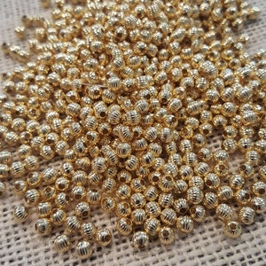 May include: A large pile of gold-colored round beads with a textured surface. The beads are scattered on a beige fabric background.