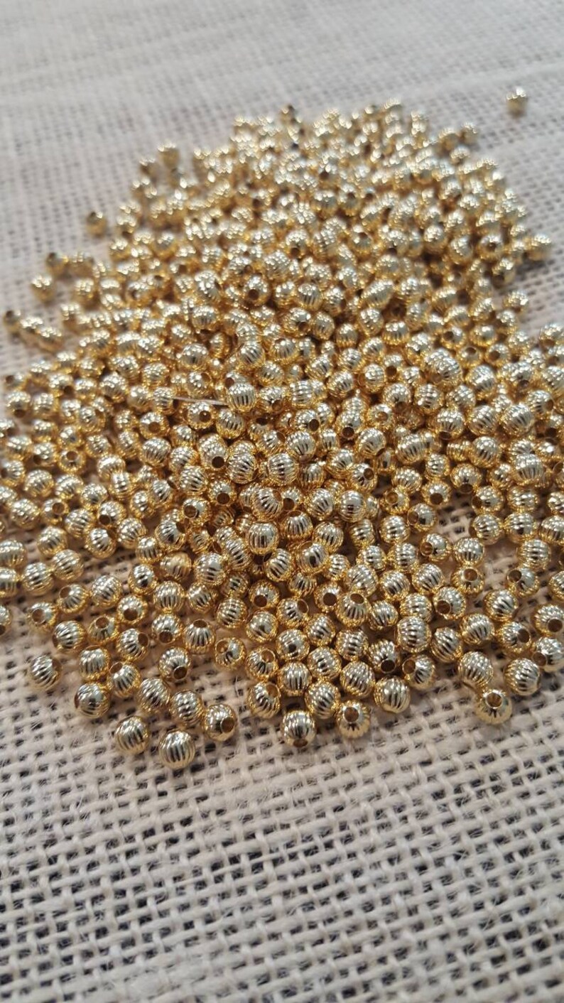 May include: A large pile of gold-colored round beads with a textured surface. The beads are scattered on a beige fabric background.