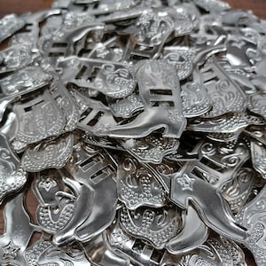 May include: A pile of shiny silver-colored metal belt buckles with a Western theme. The buckles feature designs of cowboy boots, stars, and decorative scrollwork. The buckles are on a wooden surface.