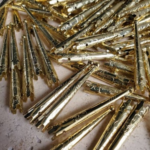 May include: A pile of gold-colored metal tips with a decorative floral pattern. The tips are pointed at one end and have a small ball at the other end.