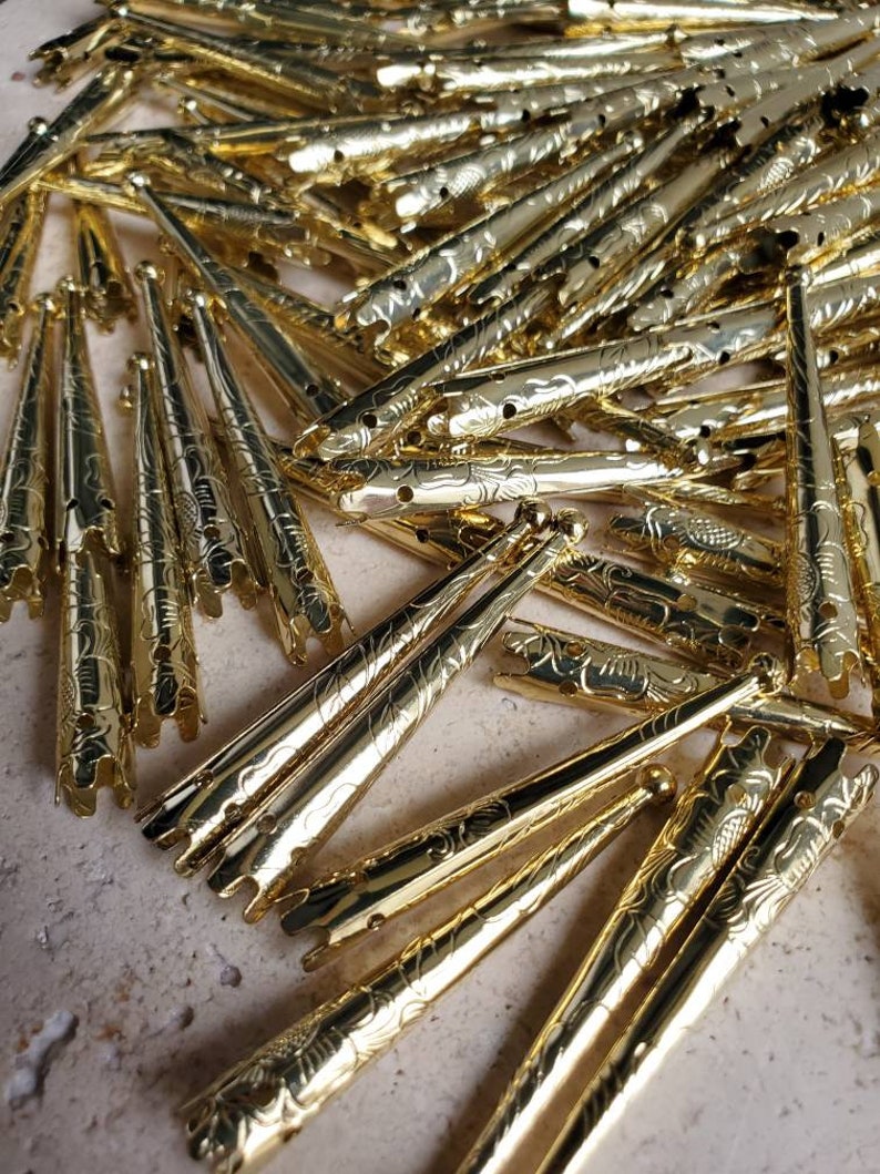 May include: A pile of gold-colored metal tips with a decorative floral pattern. The tips are pointed at one end and have a small ball at the other end.