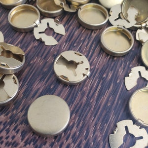 Puede incluir: Una colección de pequeños componentes metálicos de color dorado. Los componentes incluyen discos redondos y planos y piezas circulares con un centro empotrado y un borde dentado. Algunas piezas tienen un diseño en forma de estrella. Los artículos están dispersos sobre una superficie de vetas de madera oscura.