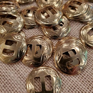 May include: A close-up of a pile of gold-colored metal buttons with a decorative design and a rectangular cutout in the center. The buttons are arranged in a random pattern on a beige fabric surface.