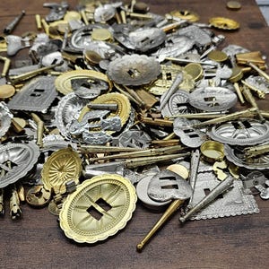 May include: A collection of silver and gold-toned metal belt buckles and decorative hardware. The assortment includes oval, rectangular, and circular shapes with various embossed designs. Some pieces have intricate patterns and cut-out details, creating a vintage aesthetic.