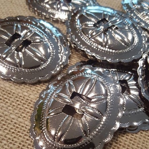 Puede incluir: Un conjunto de ocho conchos de metal plateados con un diseño floral decorativo y un agujero central. Los conchos tienen un borde festoneado y están dispuestos en una pila.