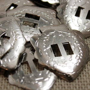 以下が含まれることがあります： 複雑なデザインが施された銀色の金属製のバックルが積み重ねられています。各バックルには、長方形の切り抜きが2つあります。バックルは、茶色の布地の背景に配置されています。