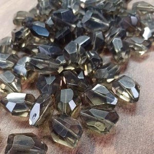 May include: A collection of faceted, smoky quartz gemstone beads. The beads have a geometric, irregular shape with multiple reflective surfaces. The colour is a translucent, smoky grey with hints of brown. The beads are scattered on a textured, brown surface.