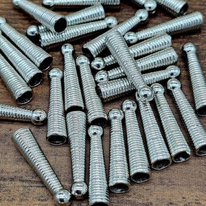 Op de afbeelding: Een verzameling zilverkleurige metalen krimpeindkralen, met een geribbeld ontwerp en een afgerond uiteinde. Deze sieradenonderdelen liggen verspreid op een houten oppervlak.