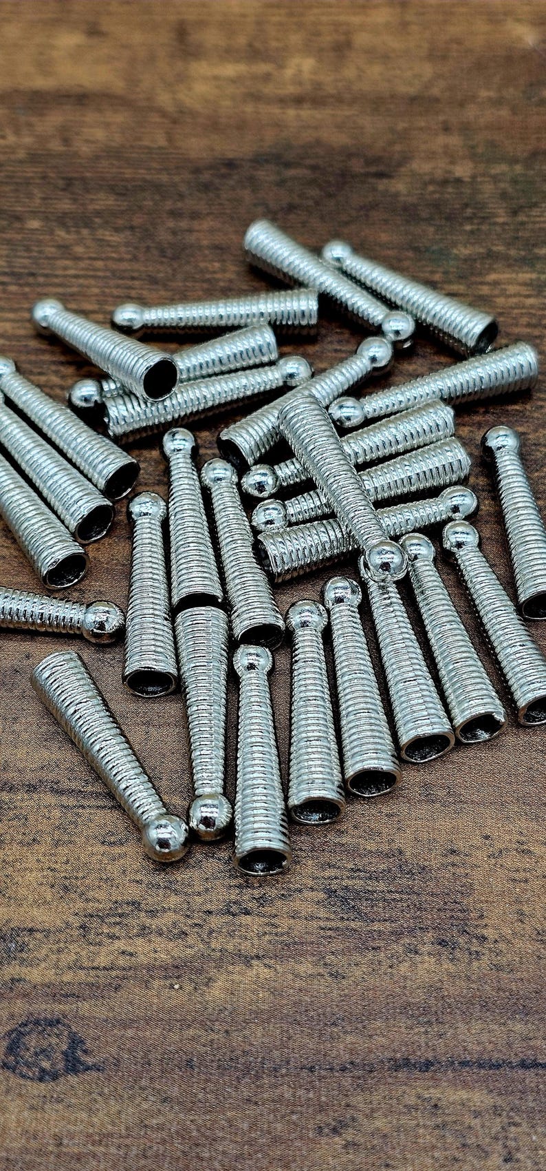 Op de afbeelding: Een verzameling zilverkleurige metalen krimpeindkralen, met een geribbeld ontwerp en een afgerond uiteinde. Deze sieradenonderdelen liggen verspreid op een houten oppervlak.