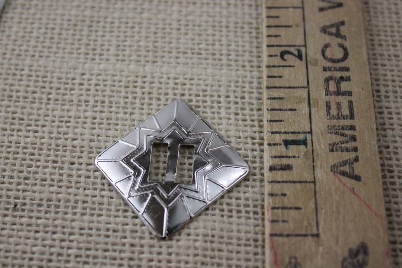 Pu&ograve; includere: Una fibbia in metallo argentato con un design a stella e un'apertura rettangolare. La fibbia misura circa 2,5 cm.