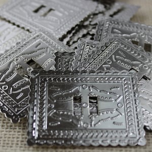 May include: A pile of silver-toned metal light switch plates with an intricate, embossed design. The plates have a square shape with rounded corners and a cutout for a light switch.