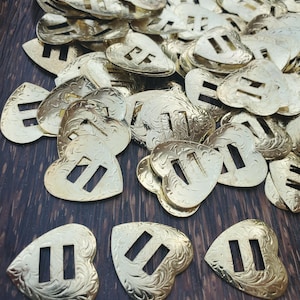 May include: A large pile of gold-coloured metal heart-shaped buckles with a decorative floral pattern and two parallel slots.