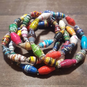 Op de afbeelding: Een kleurrijke ketting gemaakt van gerolde papier kralen. De kralen zijn in verschillende kleuren, waaronder rood, geel, blauw, groen en zwart. De ketting is gelaagd en lijkt handgemaakt te zijn.