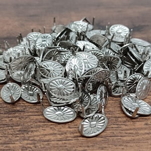 Puede incluir: Un montón de tachuelas plateadas para tapicería con un diseño decorativo en forma de sol en la cabeza. Las tachuelas tienen una cabeza redonda y plana y un pasador corto de metal. Están esparcidas sobre una superficie de madera marrón.