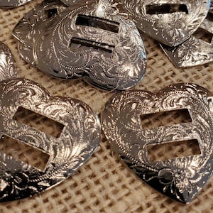 May include: Silver heart-shaped buckles with a decorative floral pattern and a double slot opening. The buckles are on a hessian background.