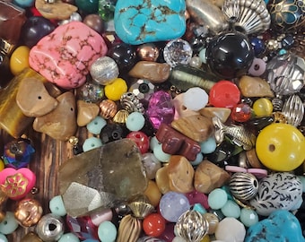 Op de afbeelding: Een verzameling kleurrijke kralen en edelstenen in verschillende vormen en maten. Bevat turquoise, roze, gele en heldere kralen, samen met metallic en iriserende accenten. Ideaal voor het maken van sieraden of knutselprojecten.