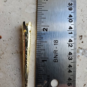 May include: A gold-colored metal stick with a decorative design and a small, round knob at the end. The stick is about 2 inches long and is measured against a ruler.