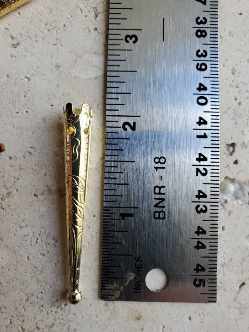 May include: A gold-colored metal stick with a decorative design and a small, round knob at the end. The stick is about 2 inches long and is measured against a ruler.