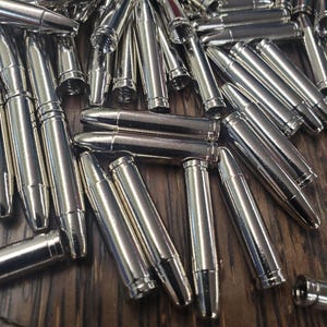 Op de afbeelding: Een verzameling glanzende, zilverkleurige hulzen verspreid over een donker houten oppervlak. De hulzen zijn cilindrisch met een puntige punt en een uitlopende basis, die licht reflecteert. Het thema suggereert munitie of een gerelateerd product.