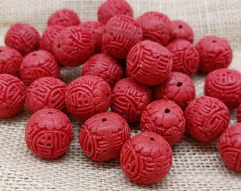 Può includere: Un insieme di perline rosse intagliate con motivi intricati. Le perline sono rotonde e hanno una finitura opaca, con un foro al centro. Sono disposte su una superficie beige strutturata.