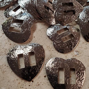 May include: A pile of silver heart-shaped metal buckles with an ornate design. The buckles have two rectangular cutouts in the centre.