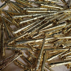 May include: A large pile of gold-colored hair clips with an ornate design. The clips are all the same size and shape, and they are arranged in a random pattern.
