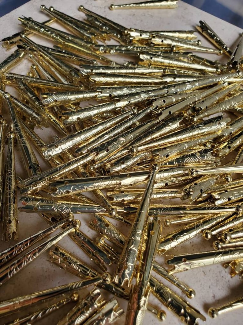 May include: A large pile of gold-colored hair clips with an ornate design. The clips are all the same size and shape, and they are arranged in a random pattern.
