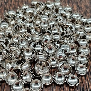 May include: A close-up of a collection of shiny, silver metal rivets. Each rivet has a rounded, domed head and a flat base with small, curved prongs. The background is a dark wooden surface.