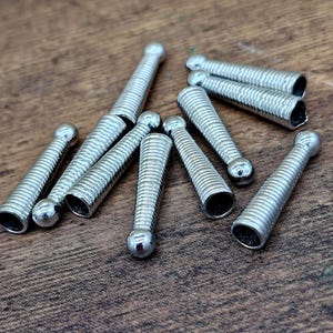 Op de afbeelding: Close-up van verschillende kleine, zilverkleurige metalen voorwerpen. Elk voorwerp heeft een afgeronde kop en een spiraalvormig lichaam. De voorwerpen liggen verspreid op een houten oppervlak.