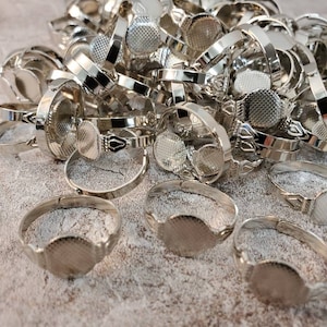 May include: A pile of silver-colored adjustable rings with oval settings. The rings have a textured surface on the setting and a simple band. Some rings have decorative details on the band.
