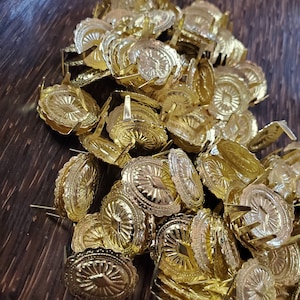 May include: A pile of gold-colored metal decorative tacks with a scalloped edge and a raised design in the center. The tacks have a pointed end for attaching to fabric or other materials.