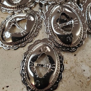 Puede incluir: Primer plano de varios conchos de metal plateados con un diseño decorativo y un recorte en el centro. Los conchos son de forma ovalada y tienen un borde festoneado.