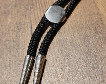 May include: A black bolo tie with a braided cord and silver-colored metal accents. The tie features a circular clasp and decorative tips. The background is a warm, wooden surface.