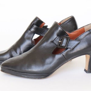 Può includere: Un paio di scarpe con tacco in pelle nera con un dettaglio fibbia e un design a taglio.