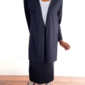 May include: A navy blue cardigan with a single button, worn over a white top and a black skirt. The outfit is completed with blue Mary Jane shoes. The clothing is simple and elegant.