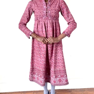 Könnte beinhalten: Ein langärmeliges rosa Kleid mit Blumendruck und weißem Kragen. Das Kleid hat einen gerafften Rock und einen Knopfleisten vorne.