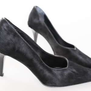 May include: A pair of black high-heeled shoes made of calf hair. The shoes have a pointed toe and a stiletto heel.